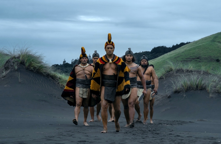 Six Māori warriors in traditional battle attire walk across black sand dunes, led by a chief at the front.