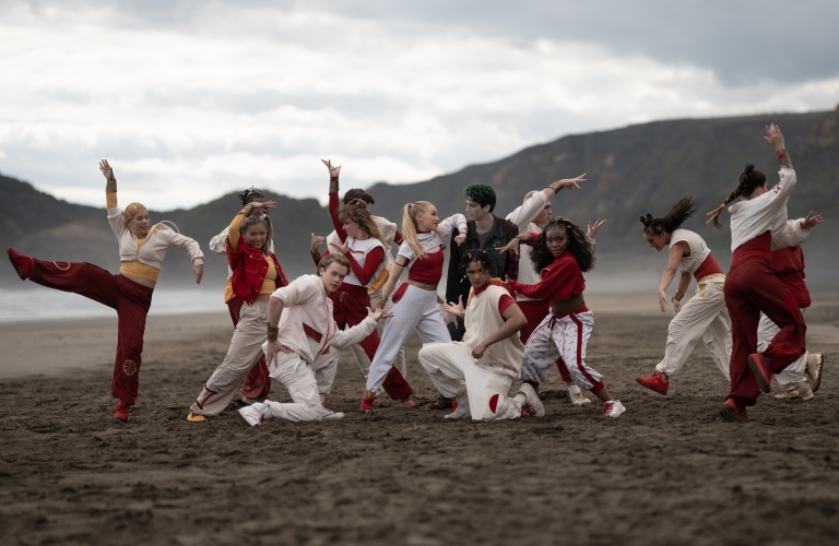 A group of dancers in red and white outfits strike energetic poses together on a black sand beach, with coastal hills in the background.