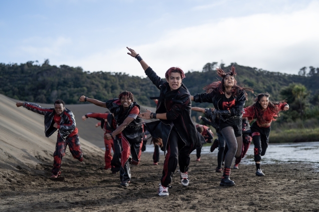 A group of dancers in black and red outfits perform an energetic routine on a black sand beach, surrounded by coastal greenery.
