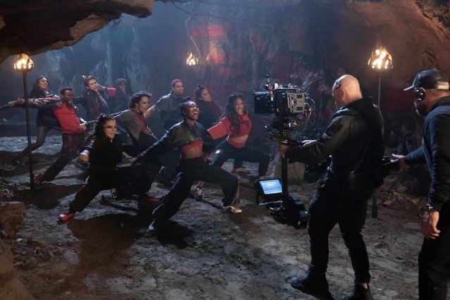 A group of dancers perform an energetic routine inside a torch-lit cave while a camera crew films the scene.